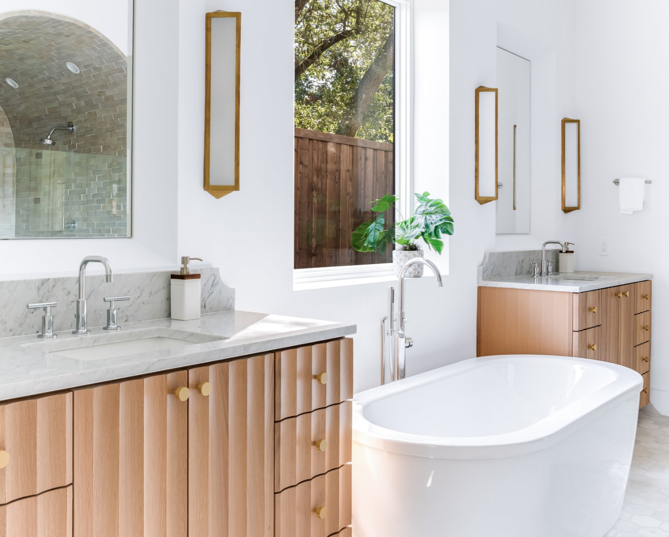 Modern bathroom with light wood vanities, a white tub, and a window overlooking greenery.