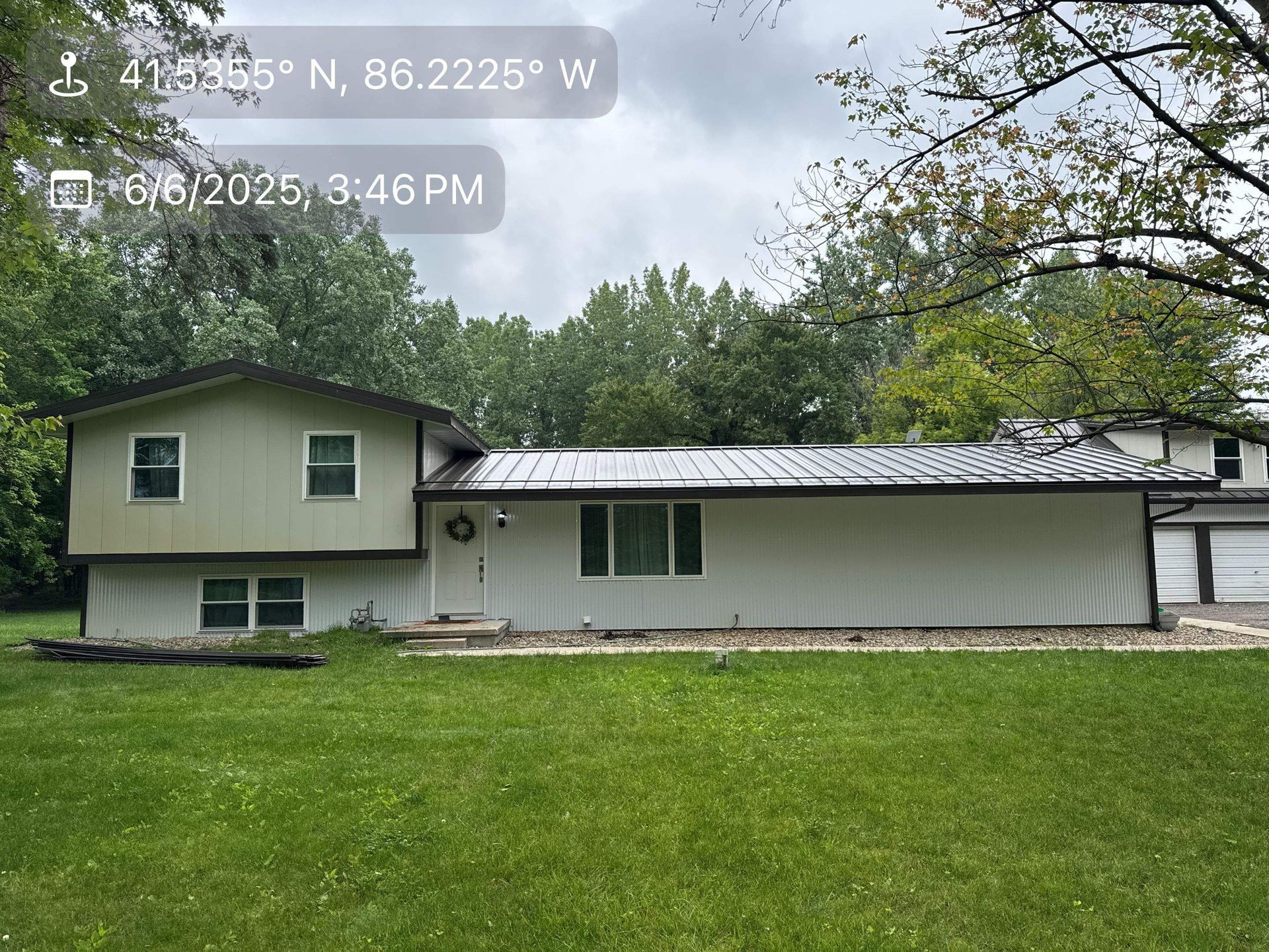 Two-story beige house with white siding and a metal roof, seen across a green lawn under cloudy trees.