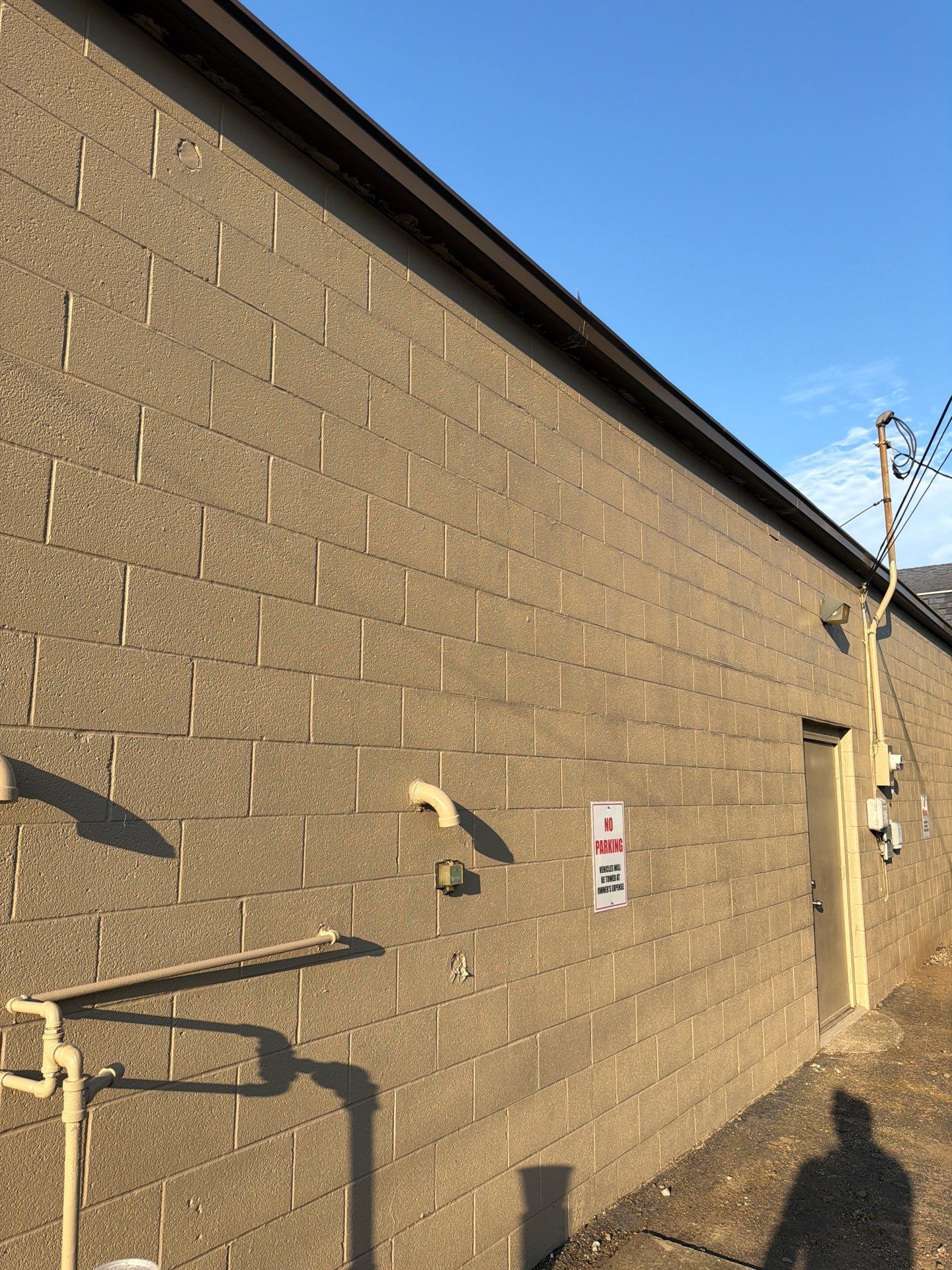 Brick building wall with mounted pipes, utility fixtures, and a shadowed sidewalk under a blue sky.