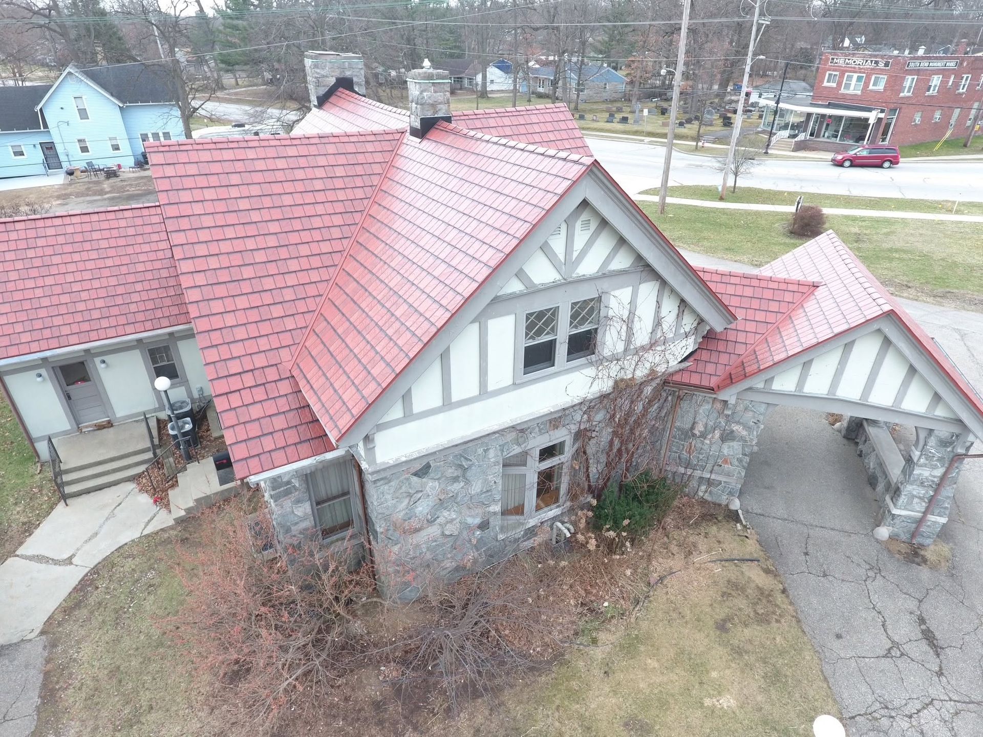Aerial view of a house with a red roof, stone front, and detached garage beside a driveway and lawn