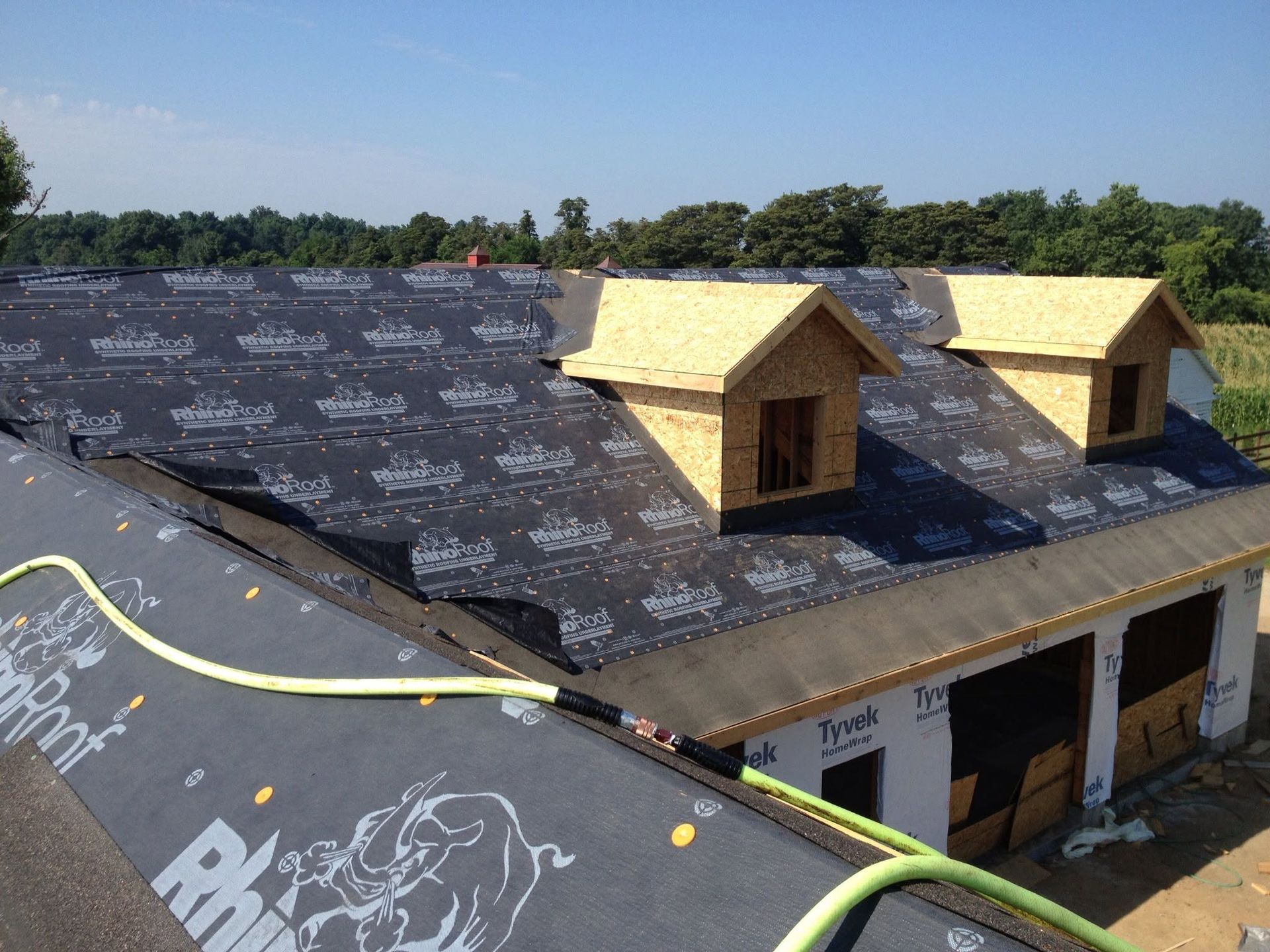 Roofing under construction with black underlayment and two dormer windows on a house under a blue sky