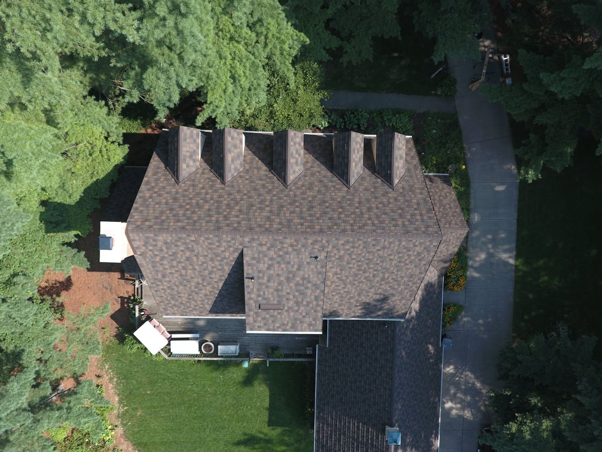 Aerial view of a house with a dark roof surrounded by dense green trees and a driveway.