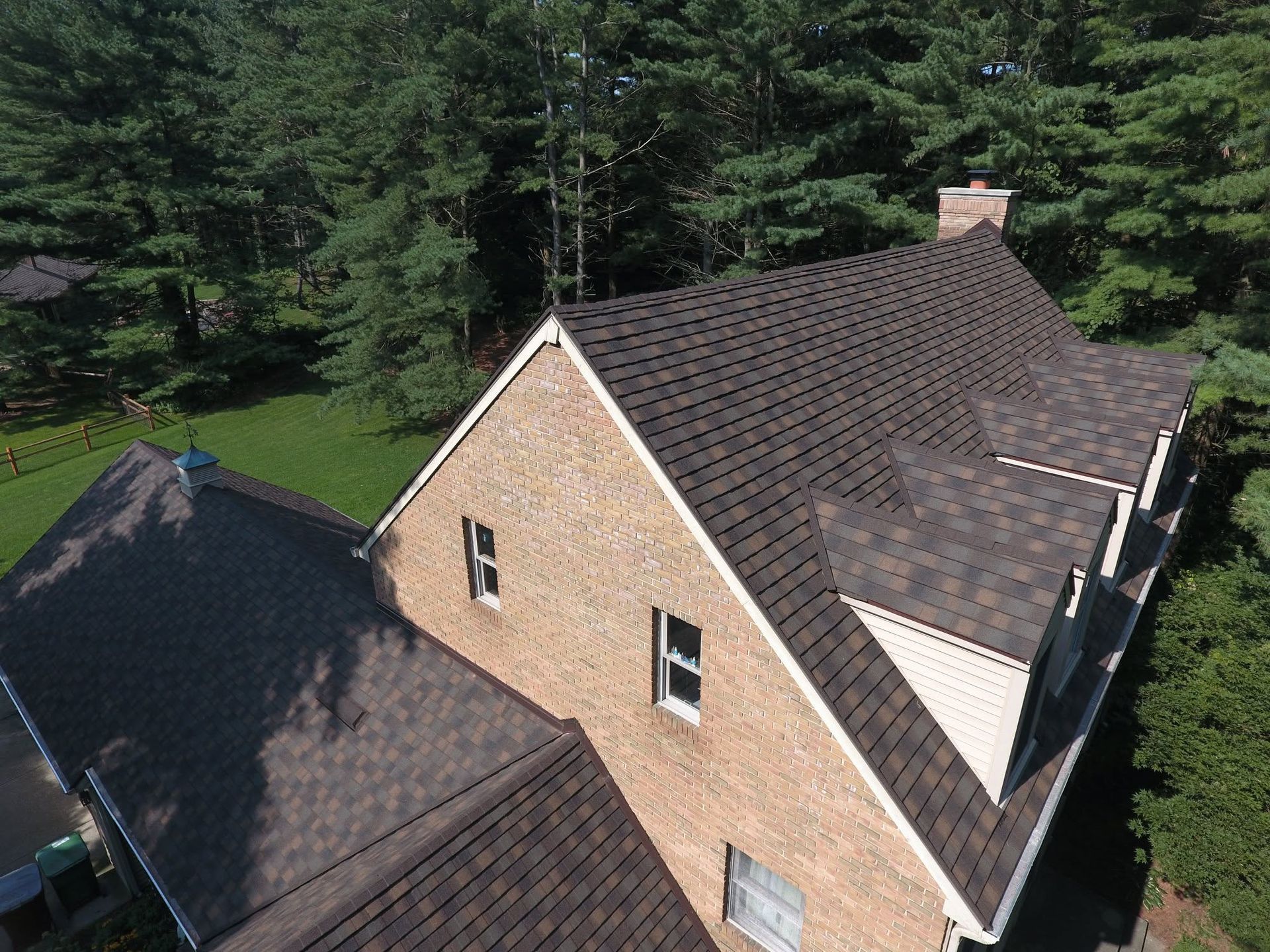 Aerial view of a brick house with dark roof surrounded by trees and lawn