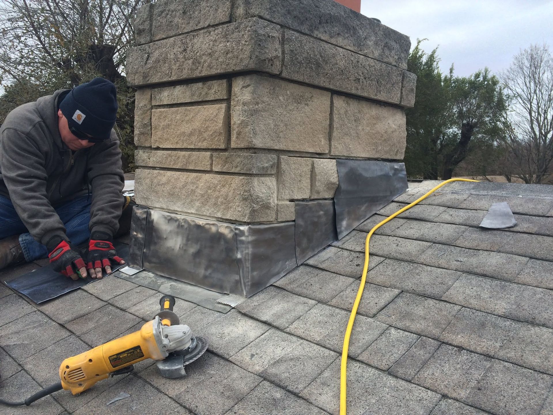 Worker repairing a stone chimney roof flashing with a yellow power tool on gray shingles
