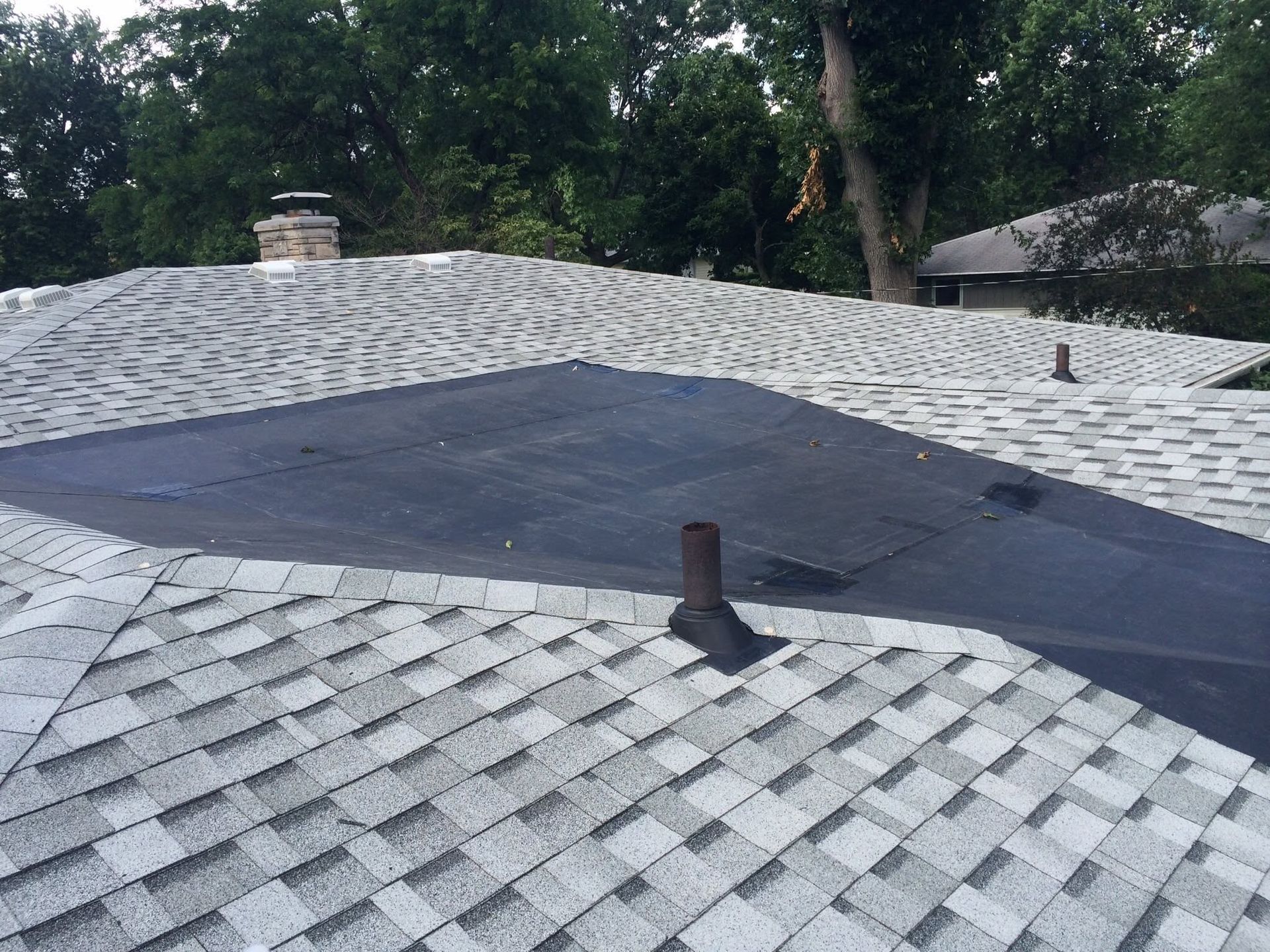 Roof with gray shingles and a large black tarp covering damaged sections beneath trees