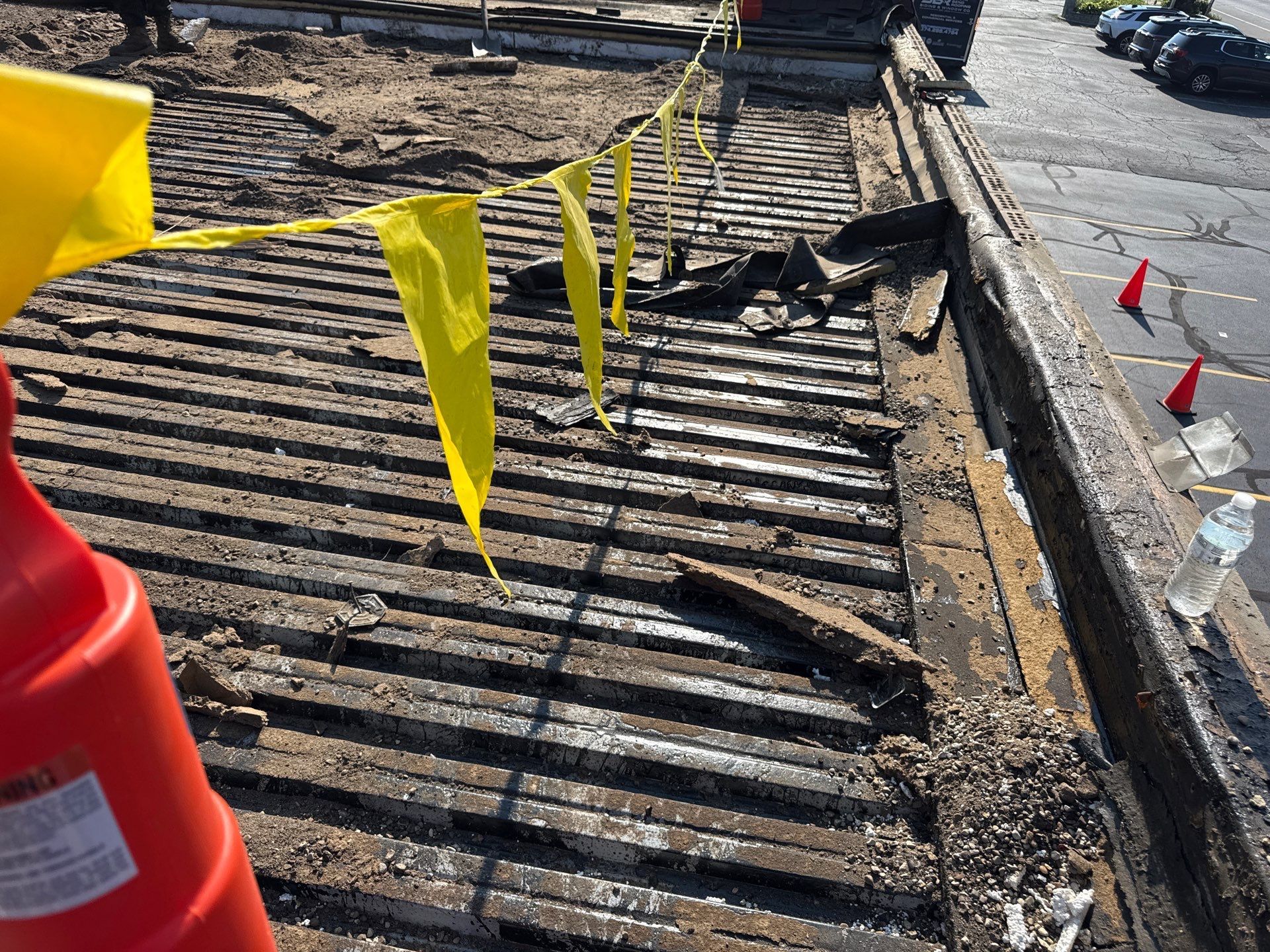 Yellow caution tape beside a damaged paved surface and curb, with an orange traffic cone in the foreground