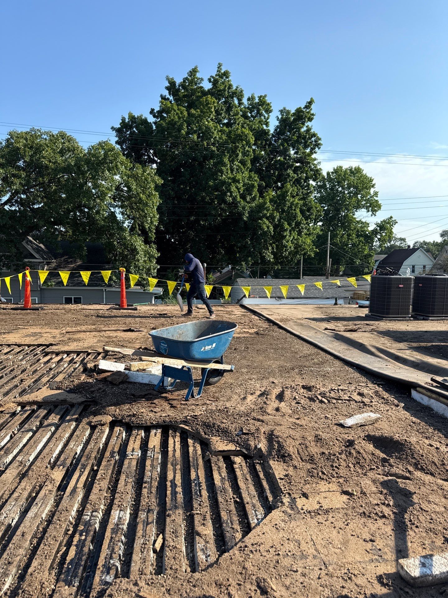 Construction site with muddy ground, pipes, and workers near a tree under a blue sky