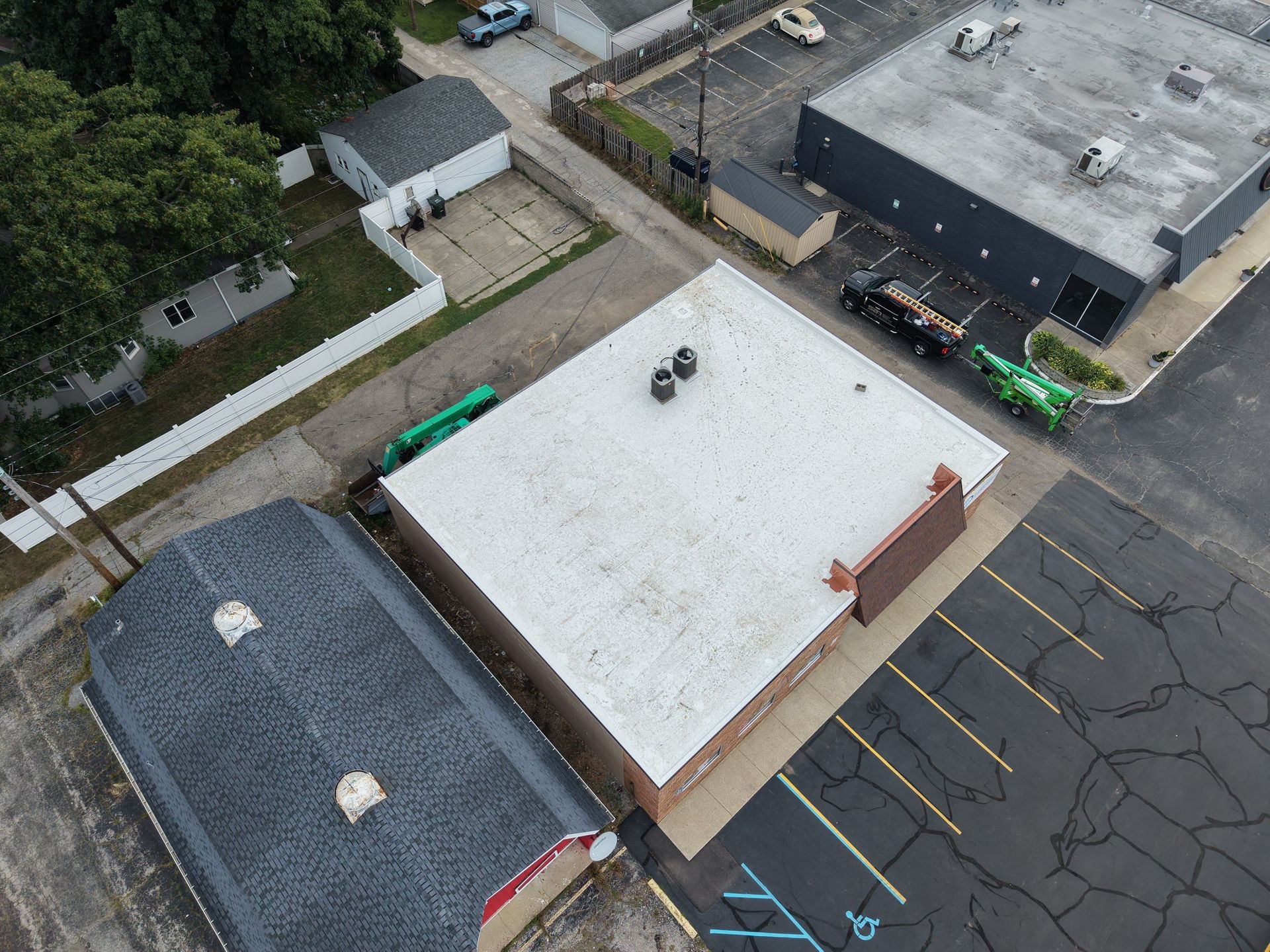 Aerial view of a flat-roofed building beside parking lots and streets, with trees and nearby structures around it.
