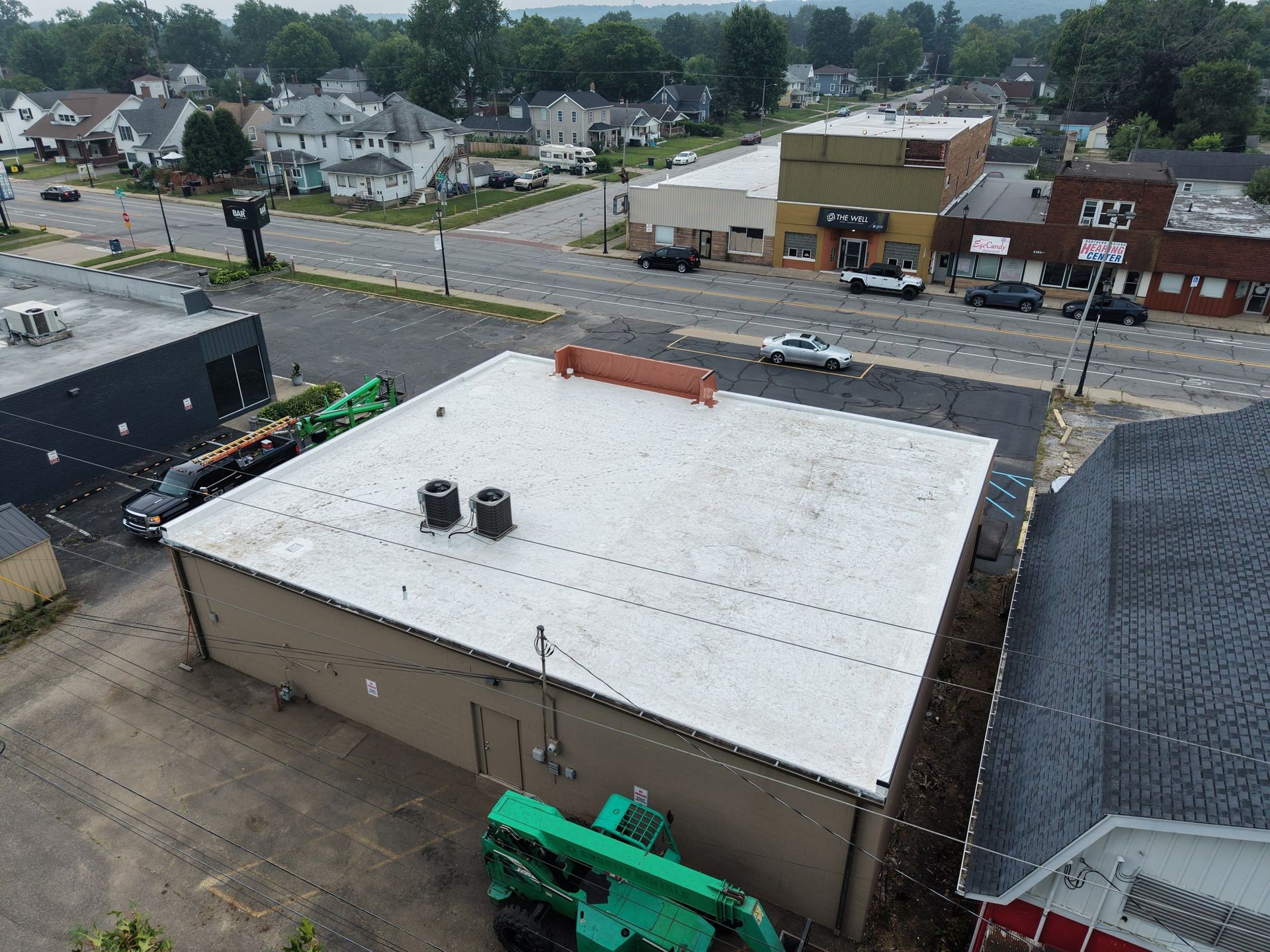 Aerial view of a large white-roofed building beside a street and nearby houses, with trucks and equipment below.