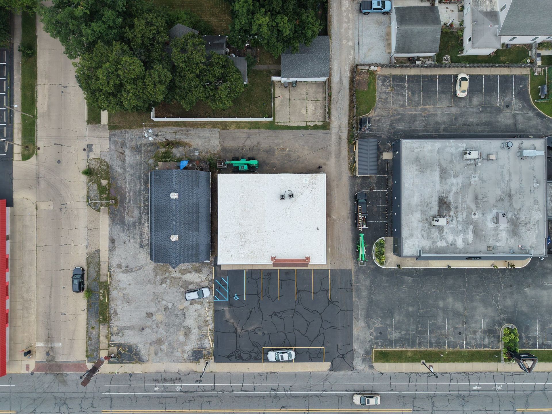 Aerial view of a small commercial lot with two flat-roof buildings, parking spaces, trees, and adjacent road.