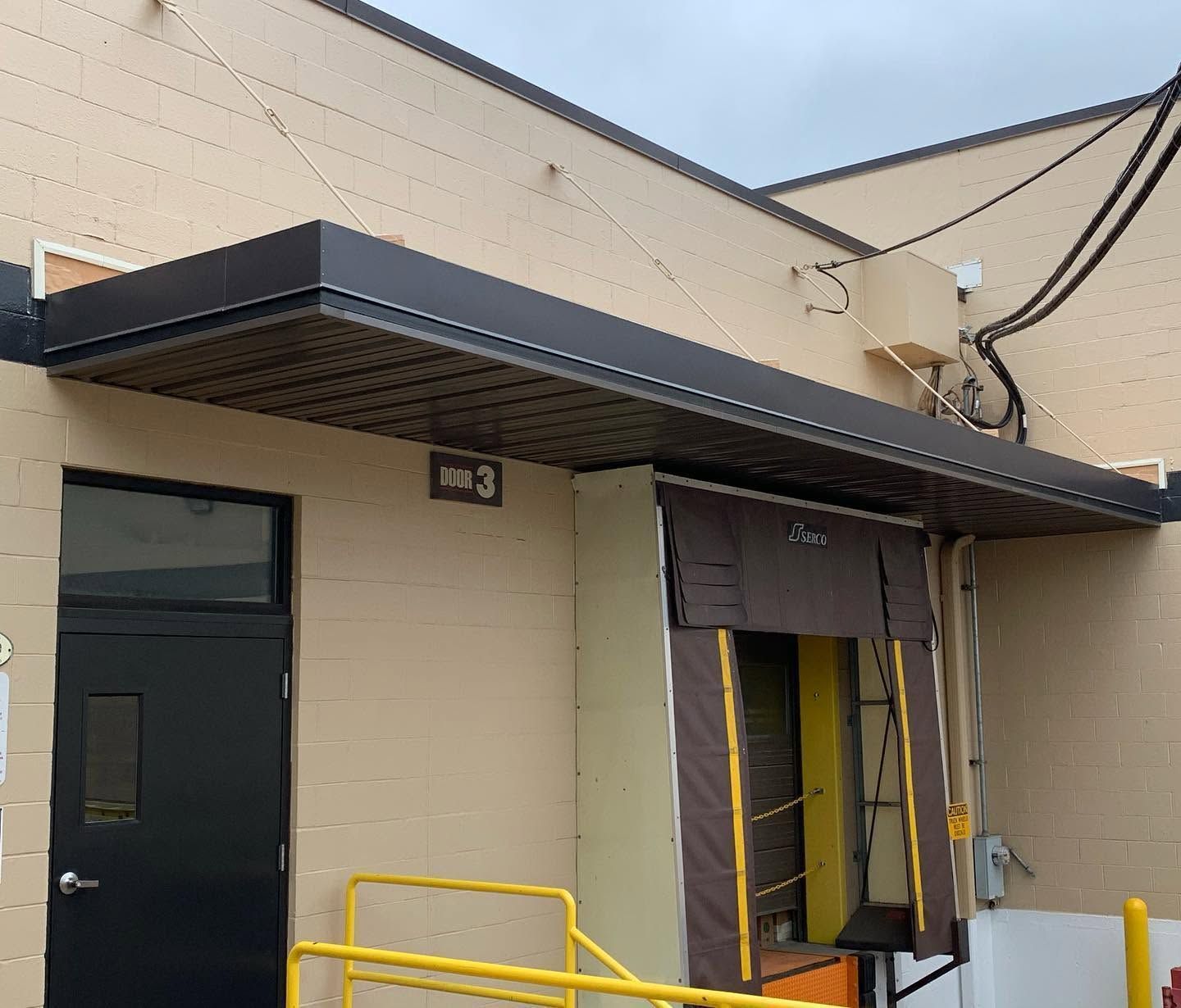 Exterior of a beige building with black awning, loading door, and yellow safety railings