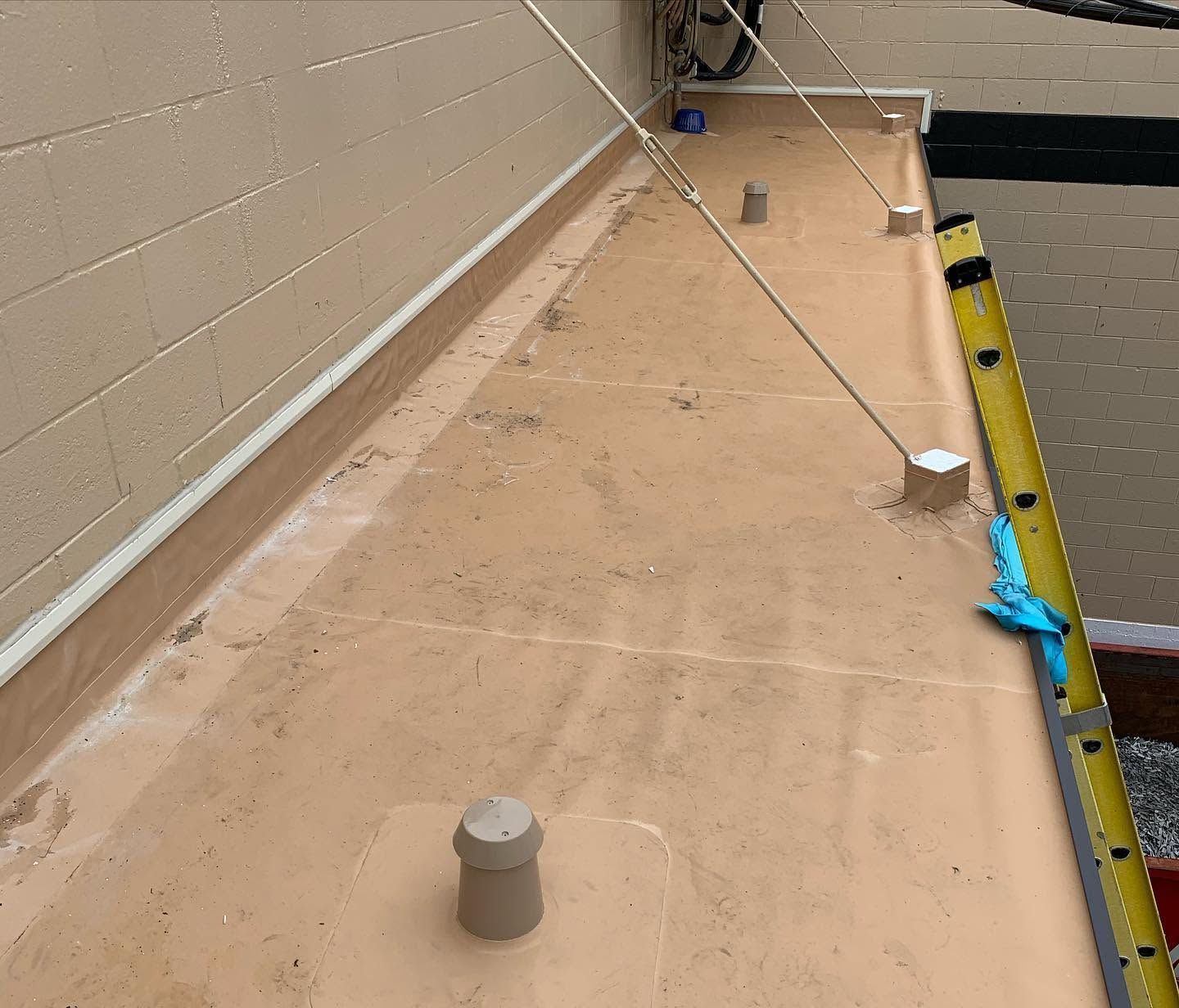 Rooftop walkway with tan membrane, wall to left, metal poles, and yellow edge barrier on right.