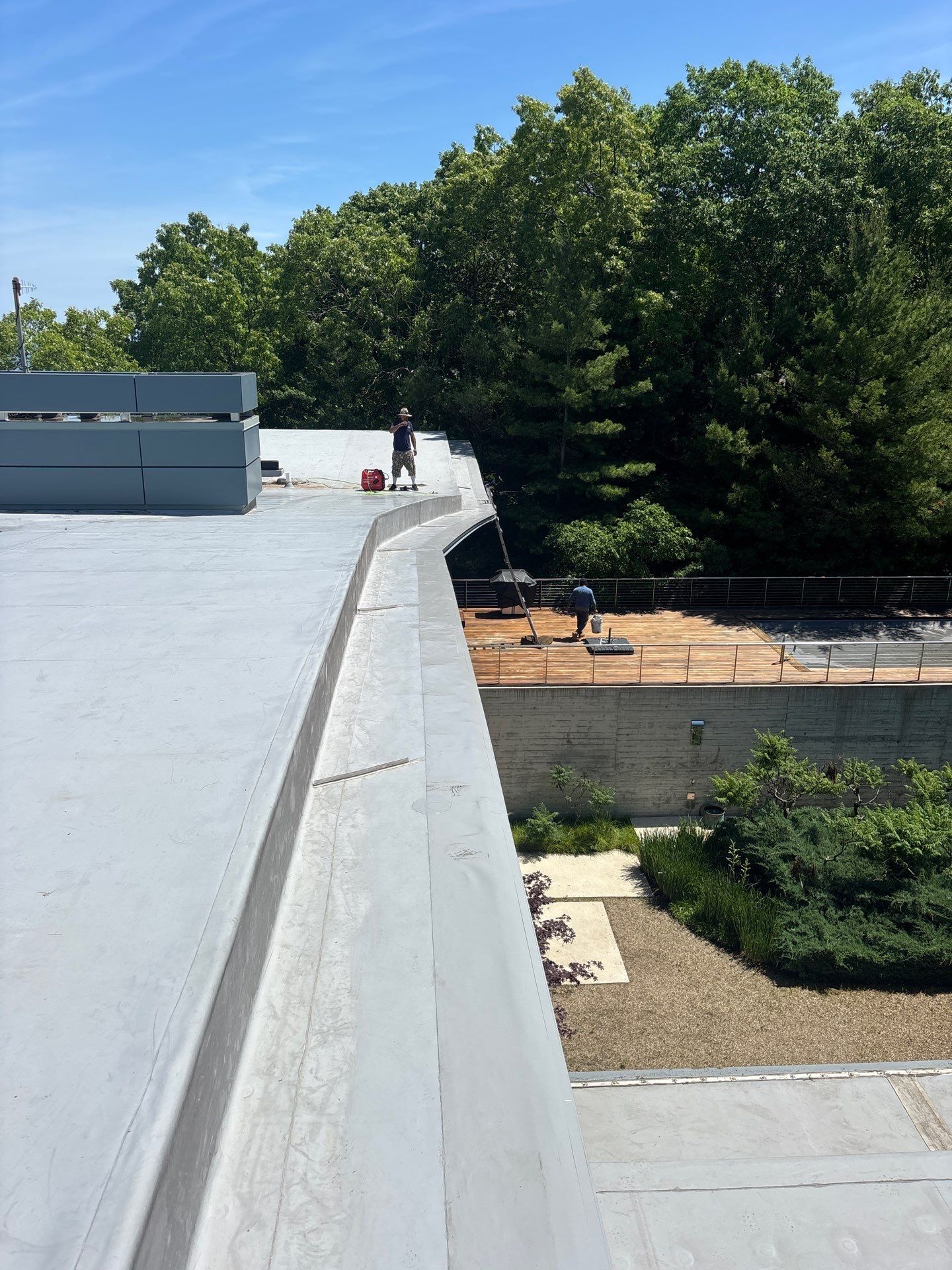 Rooftop parapet overlooking trees, a lower roof, and a distant person near a red bucket.