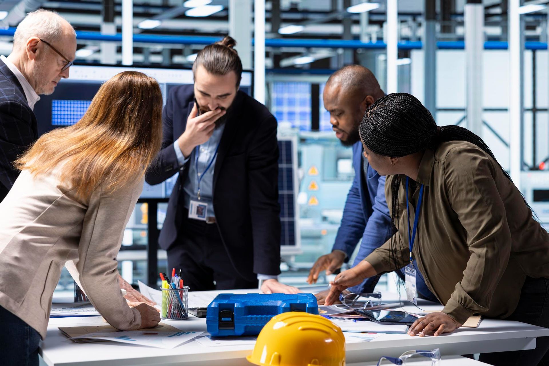 Diverse professionals collaborate around a table, reviewing documents in an industrial setting with a yellow hard hat.