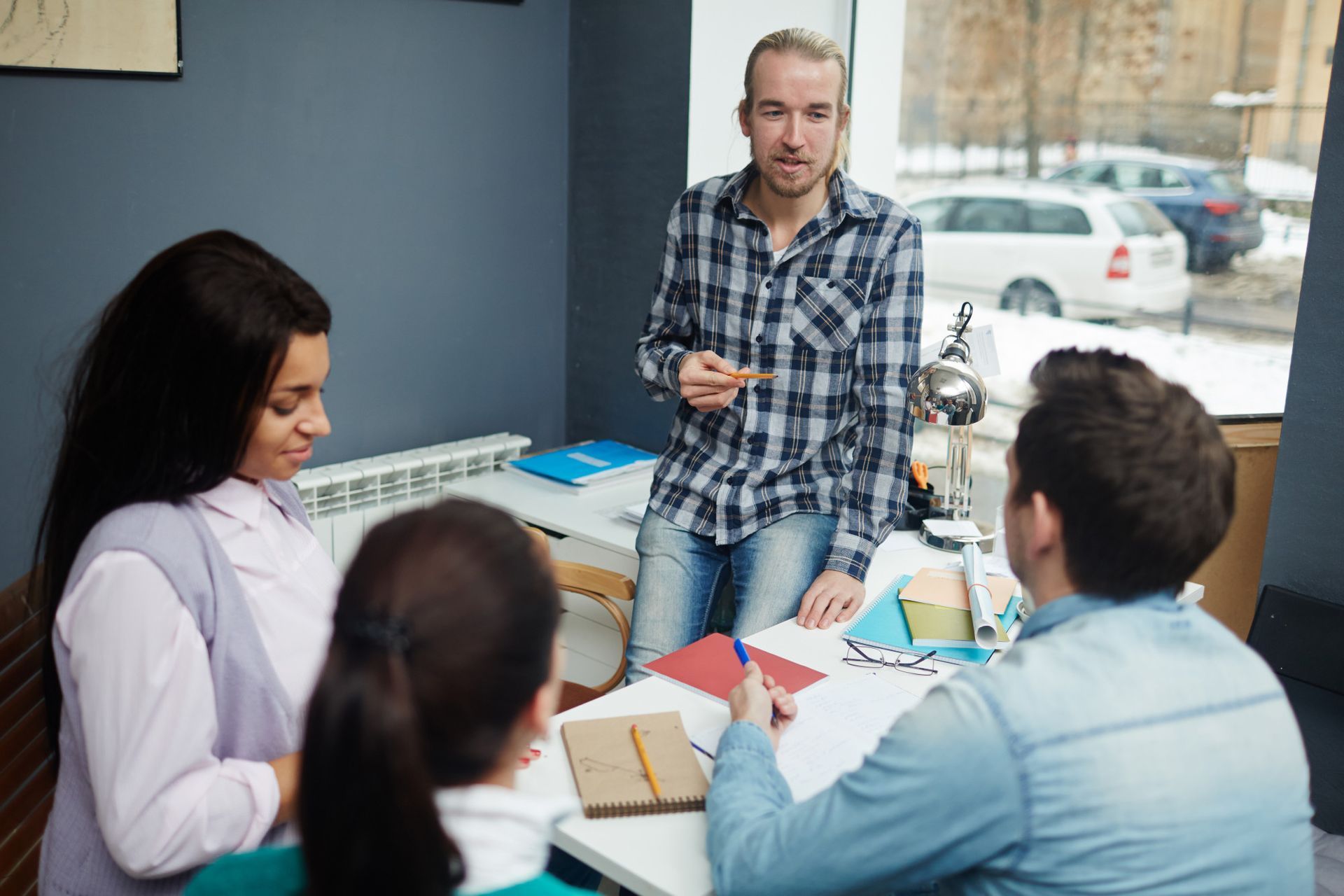 A group of four people sits around a table in an office, collaborating on a project with notebooks and documents.