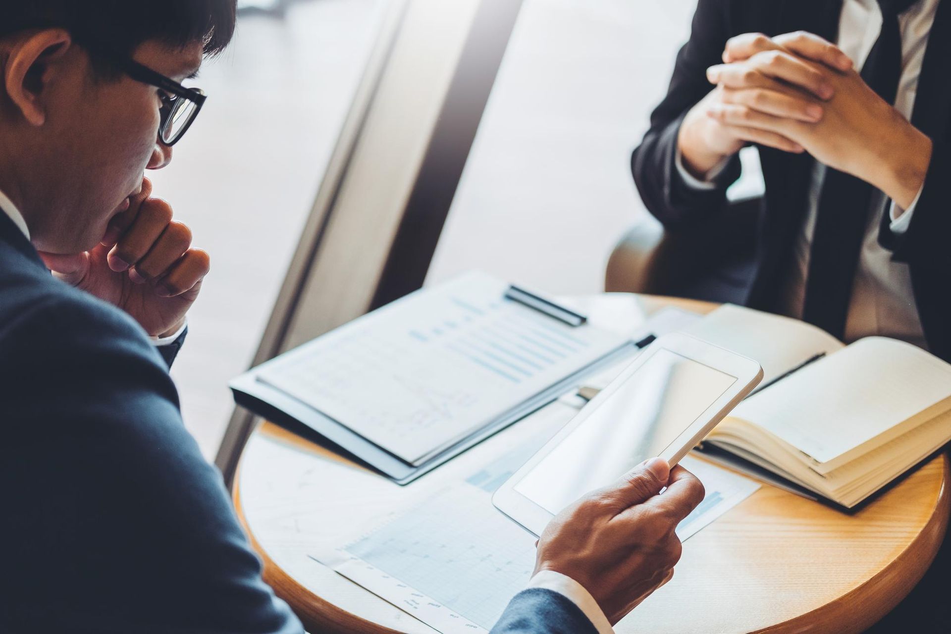 Two people in business attire discuss work at a small round table with documents and a tablet.