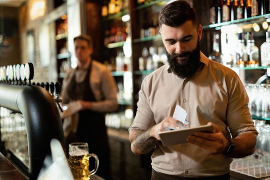 A bearded bartender in a tan shirt looks at a tablet at a bar, while a colleague works in the blurred background.