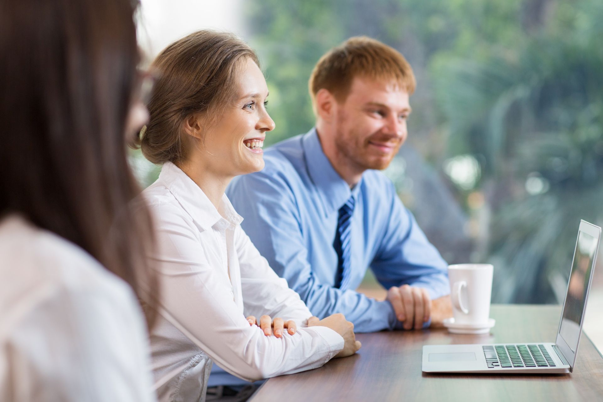 A woman and a man in business attire sit at a table smiling while looking at a laptop in a bright, modern office space.