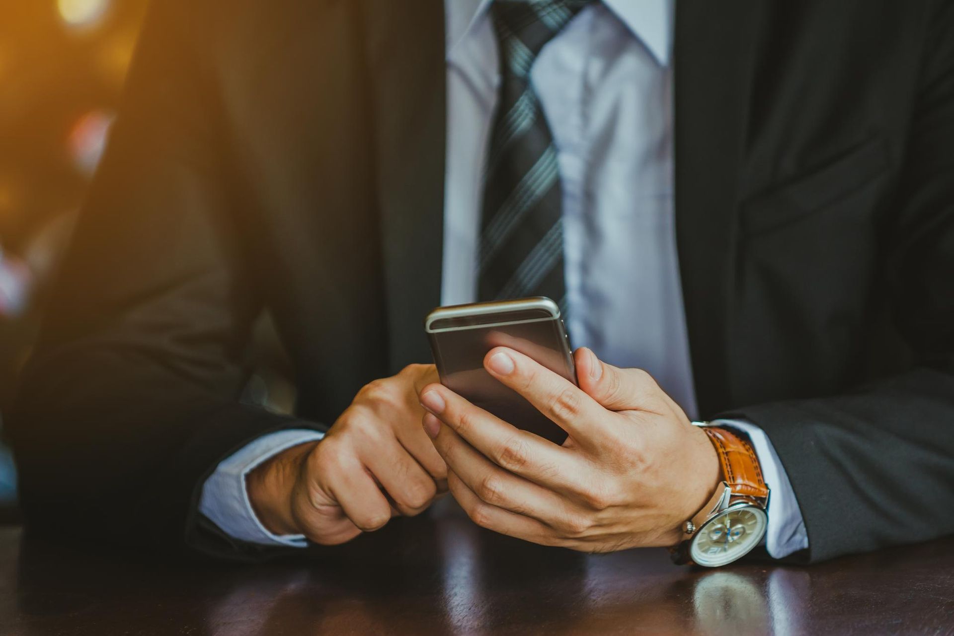 A person in a dark business suit and tie uses a smartphone while sitting at a wooden table.