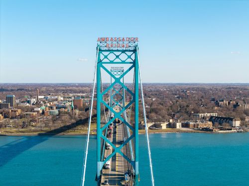 An aerial view of the turquoise, steel-truss Ambassador Bridge spanning a blue river toward a city skyline.