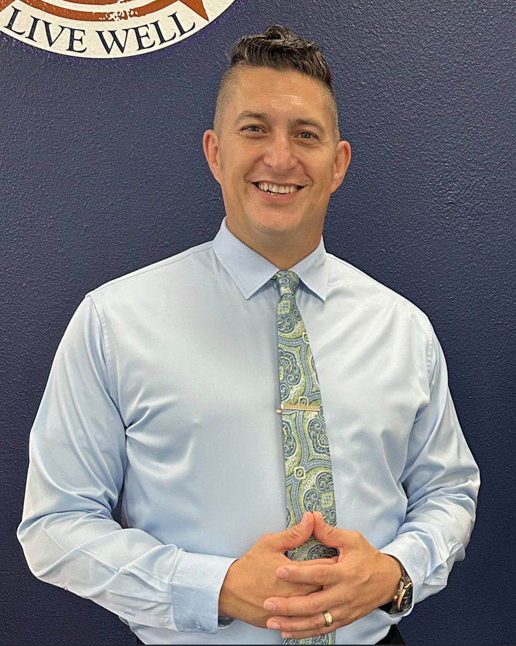 Man in light blue shirt, patterned tie, arms crossed, smiling, in front of a blue wall with a