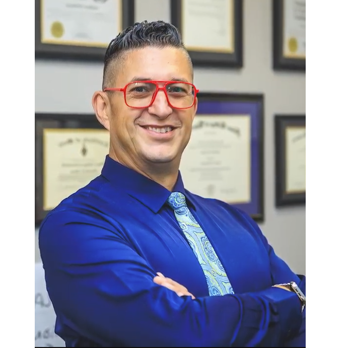 Man in blue shirt with red glasses smiling in an office, arms crossed, framed certificates behind him
