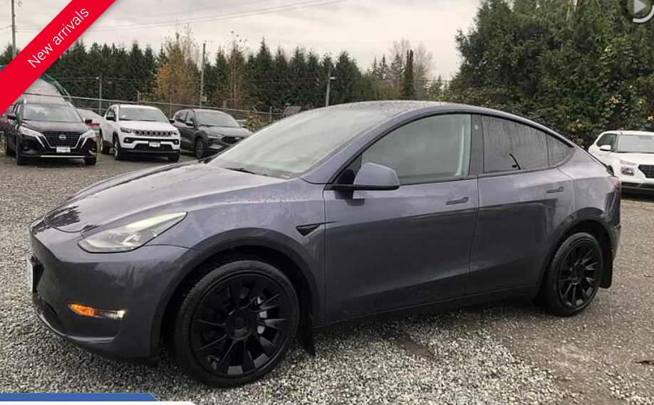 Gray Tesla Model Y parked in front of other vehicles on a gravel lot. Black rims.