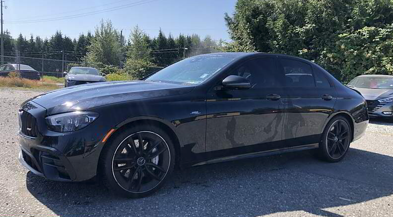 Black sedan parked on gravel in a sunny setting with trees and other vehicles in the background.