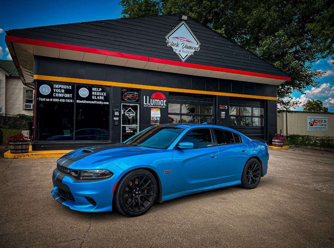 A bright blue Dodge Charger parked in front of a shop with a black exterior and a Lumar window tint logo.