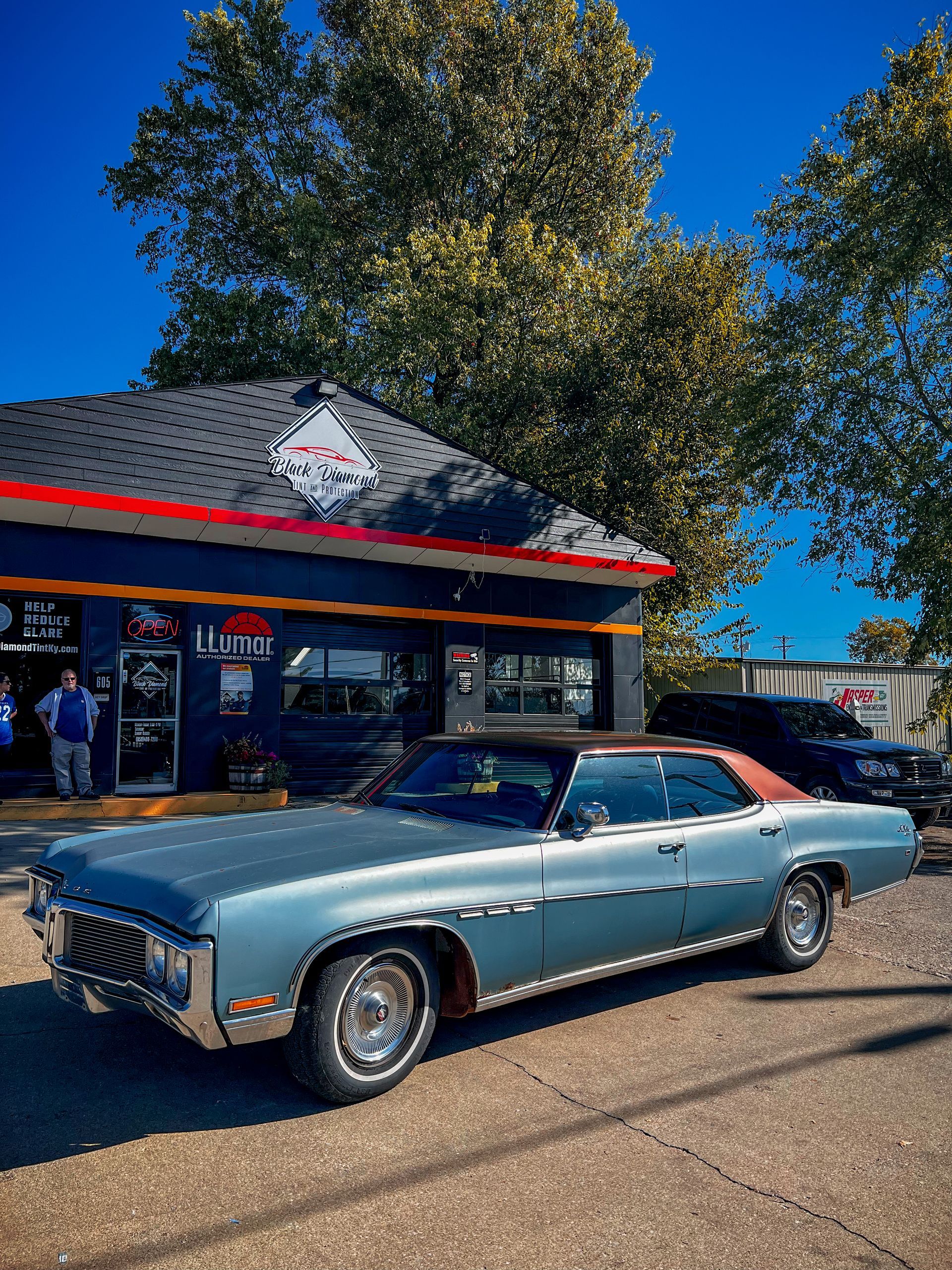 A vintage light blue Buick parked in front of a blue storefront under a clear blue sky on a sunny day.