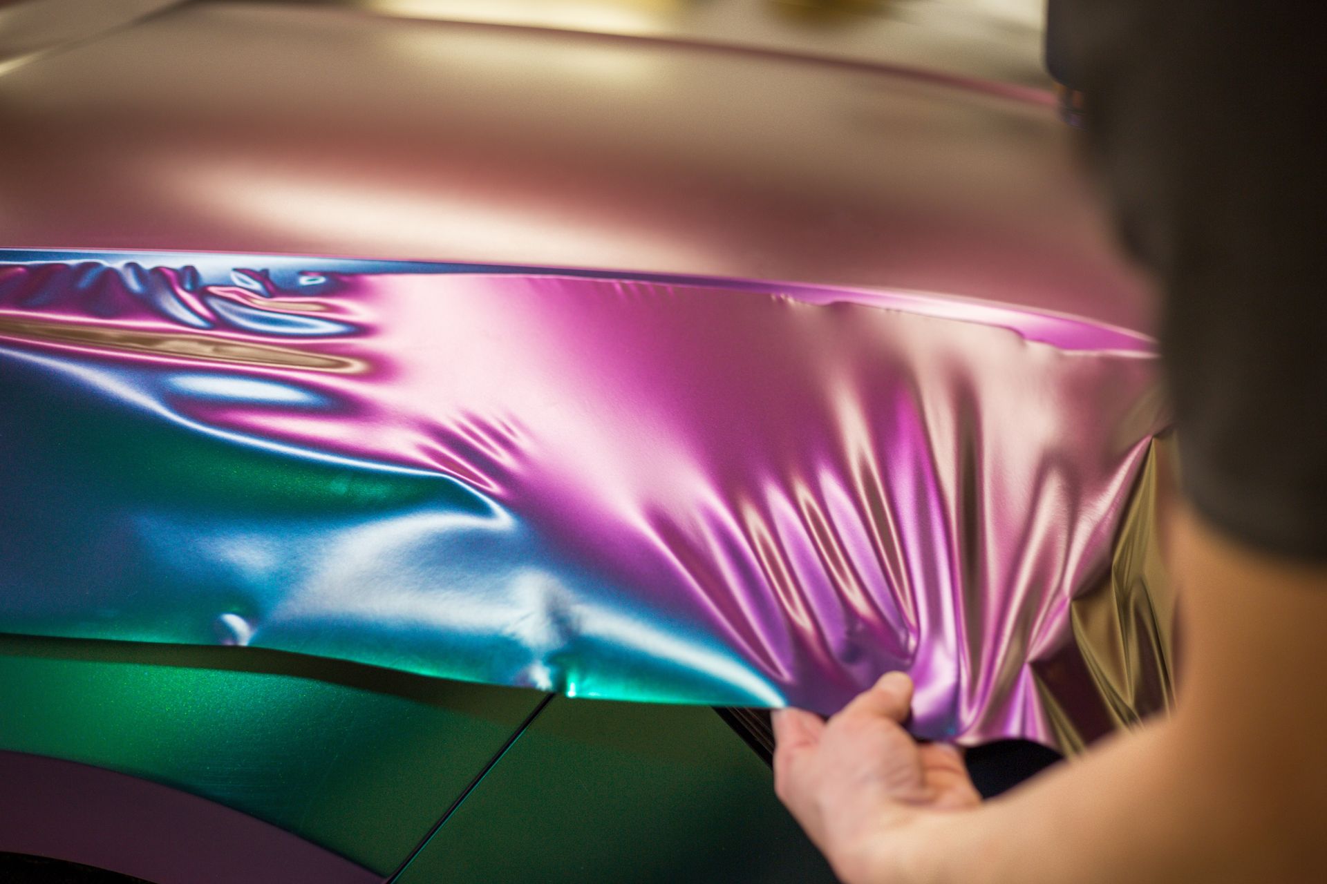 A close-up of a hand applying a color-shifting, iridescent purple and blue vinyl wrap to a car hood.