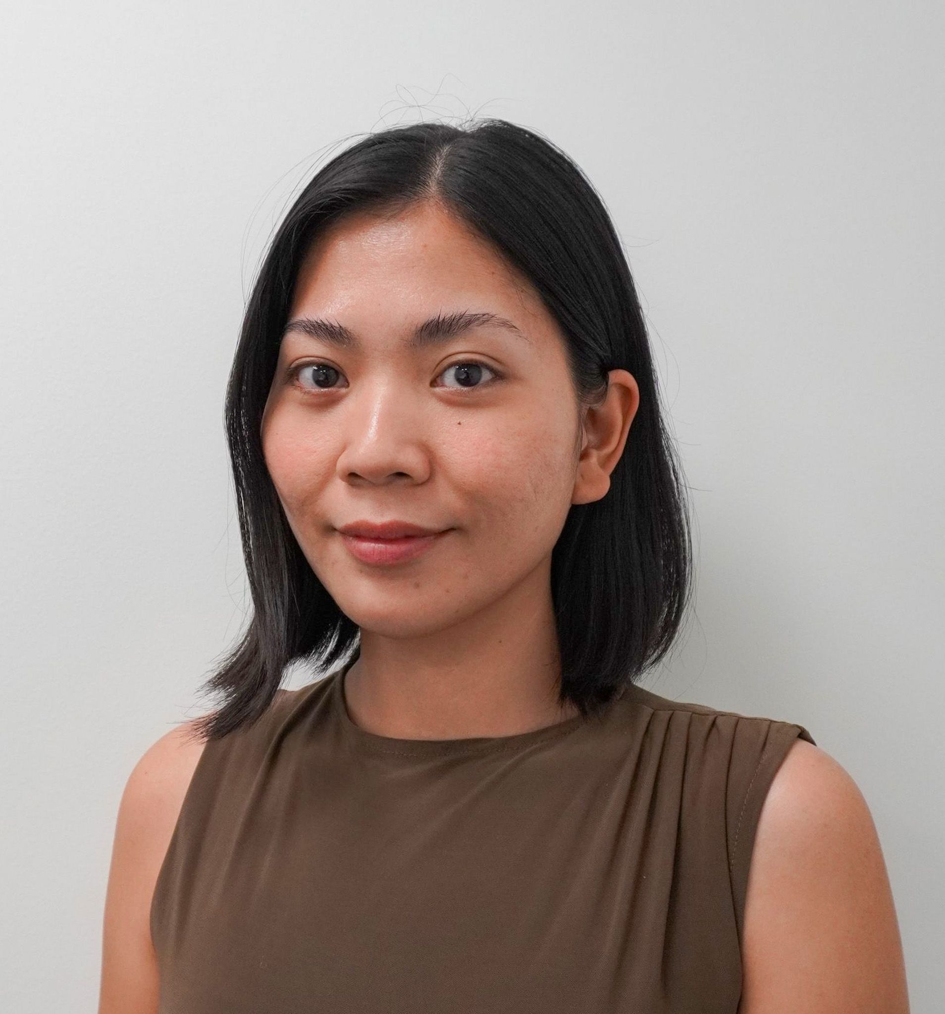 Portrait of a person with shoulder-length black hair in a brown sleeveless top against a light gray wall