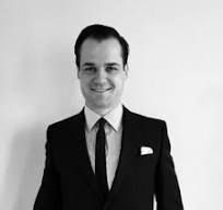 Man in a suit smiles, standing against a plain white background.