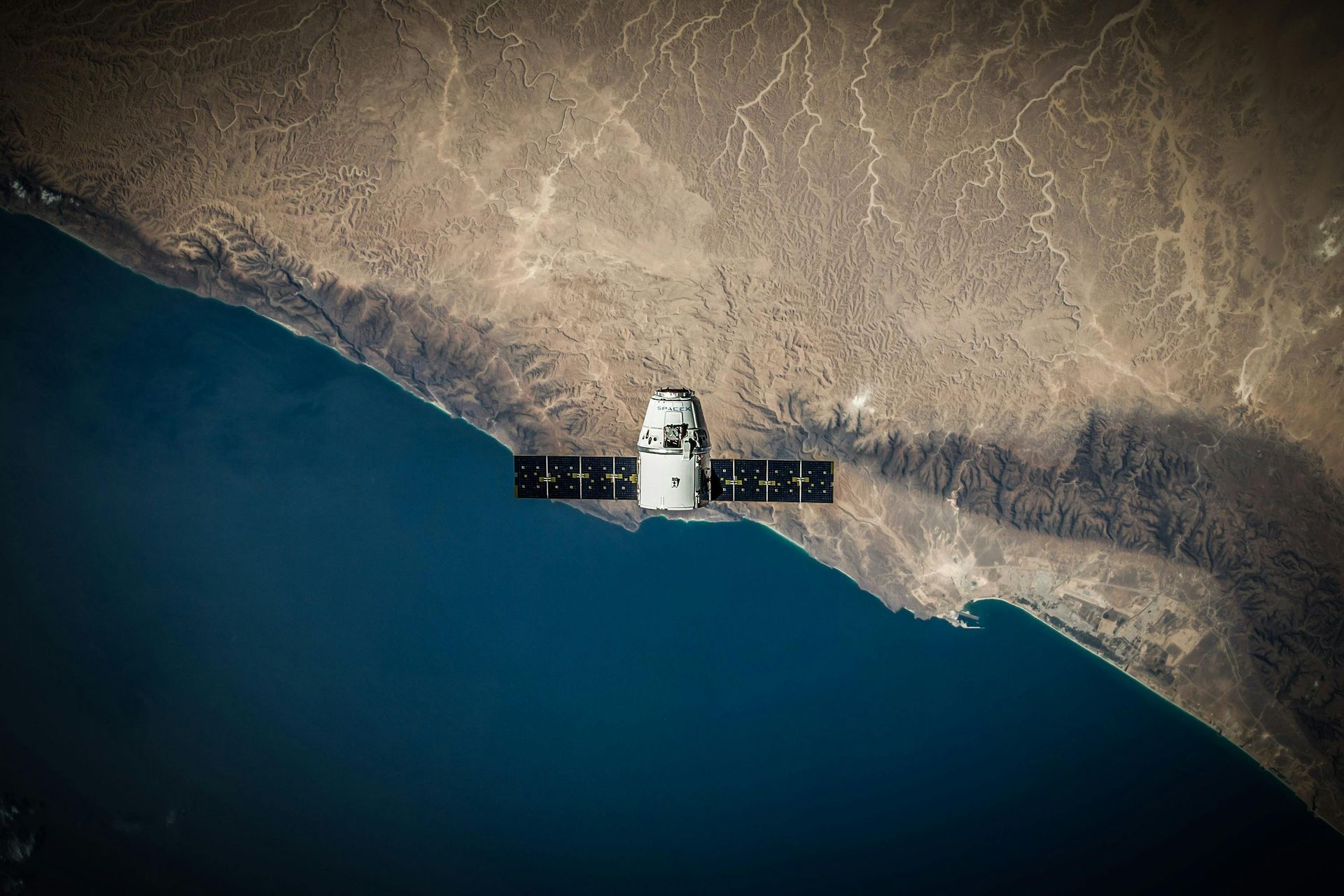 SpaceX Dragon spacecraft above coastline. Ocean and brown land with the spacecraft's solar panels extended.