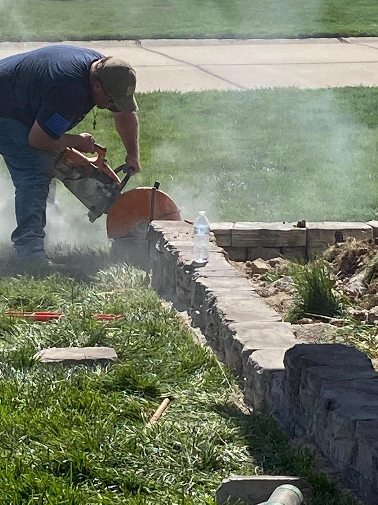 Worker using a chainsaw to cut a stone wall in a grassy yard, with dust rising.