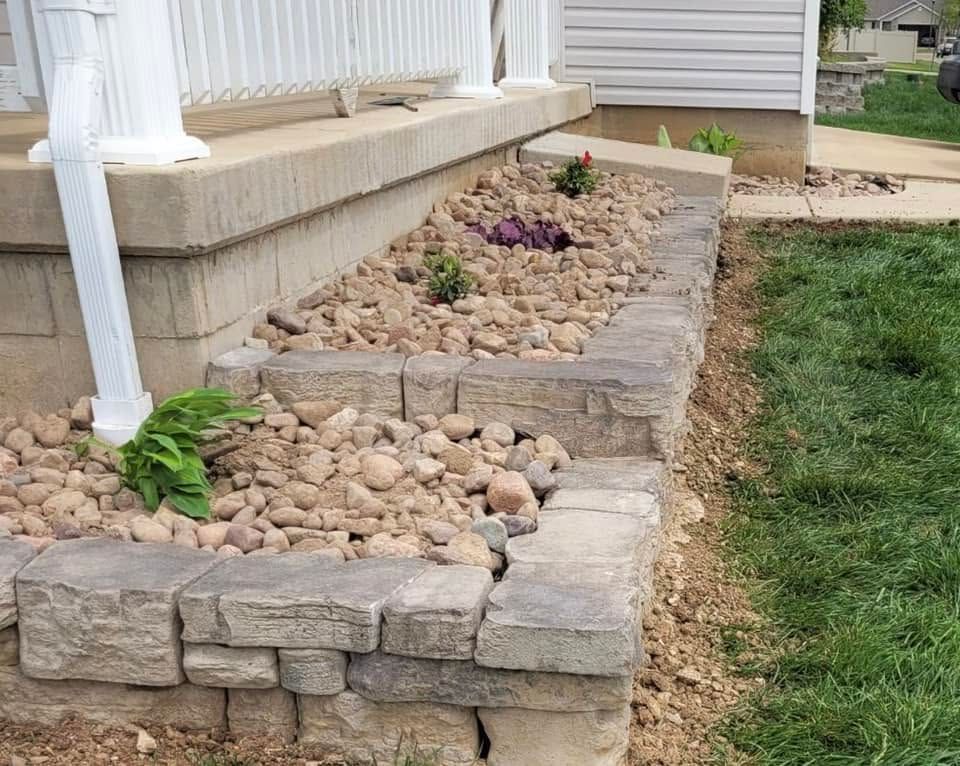 Rock-lined front yard bed with stone retaining wall and sparse plants beside a house