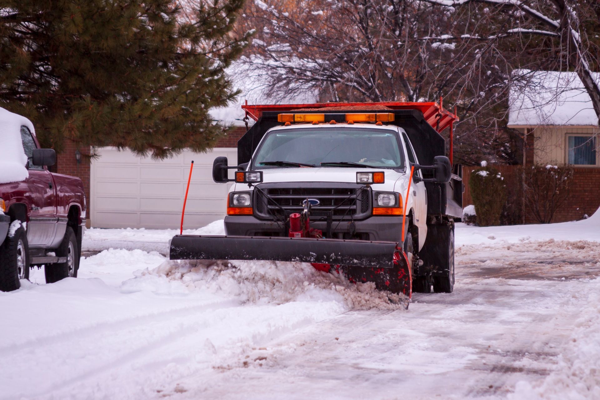 Snowplow clearing a snowy residential street, with a white truck and orange plow blade.