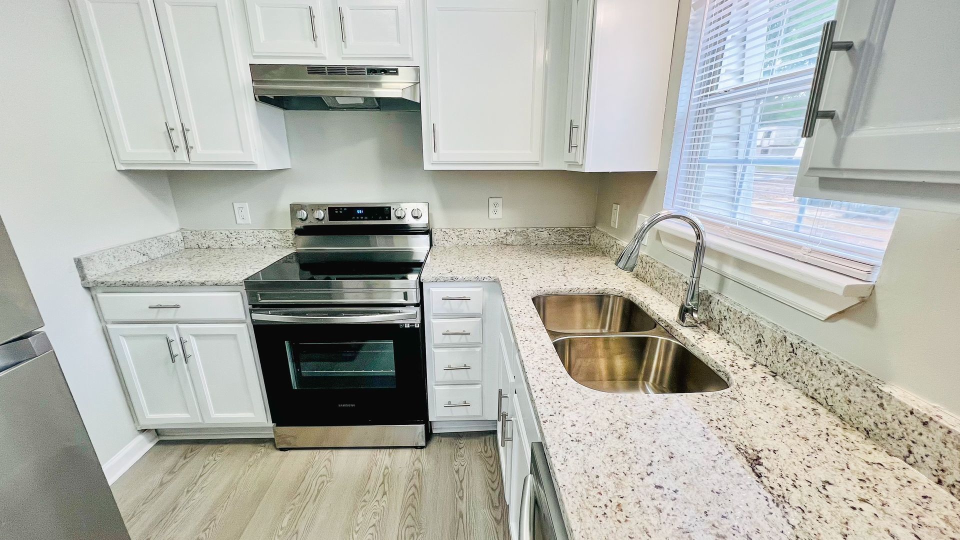 Bright white kitchen with black stove, speckled countertops, and stainless sink by a window.