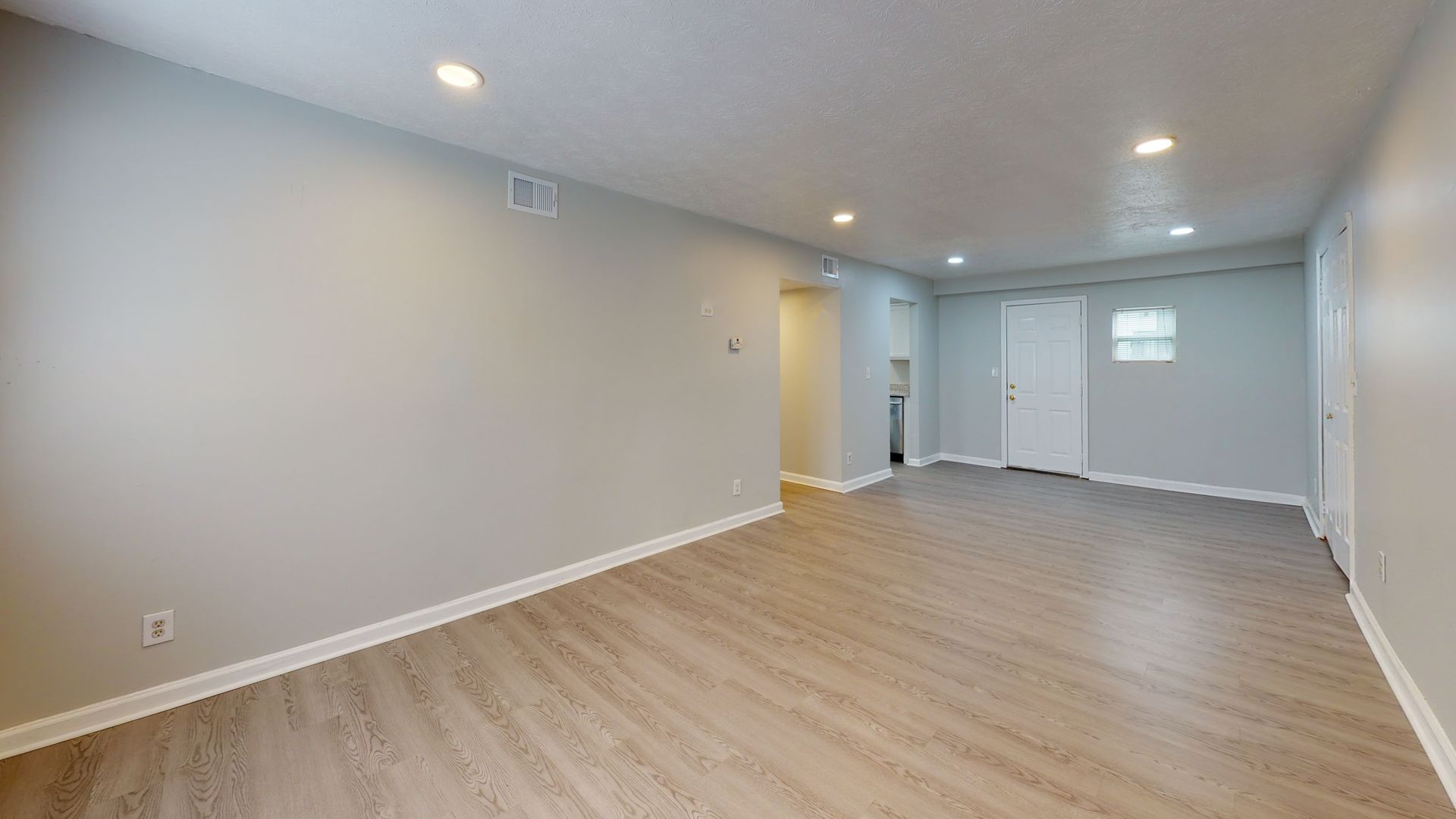 Empty modern living room with light gray walls, wood flooring, recessed lights, and a doorway in back.