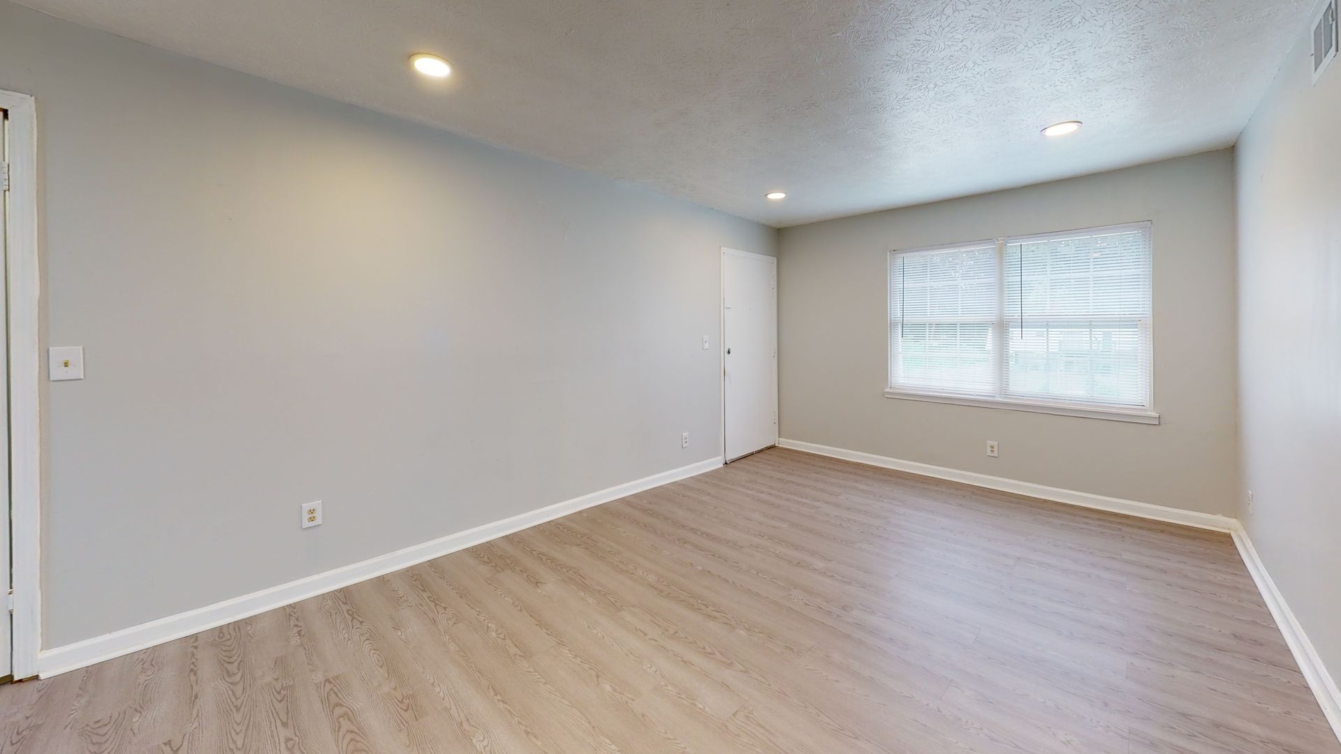 Empty light-gray room with wood floors, recessed lights, and a window with blinds