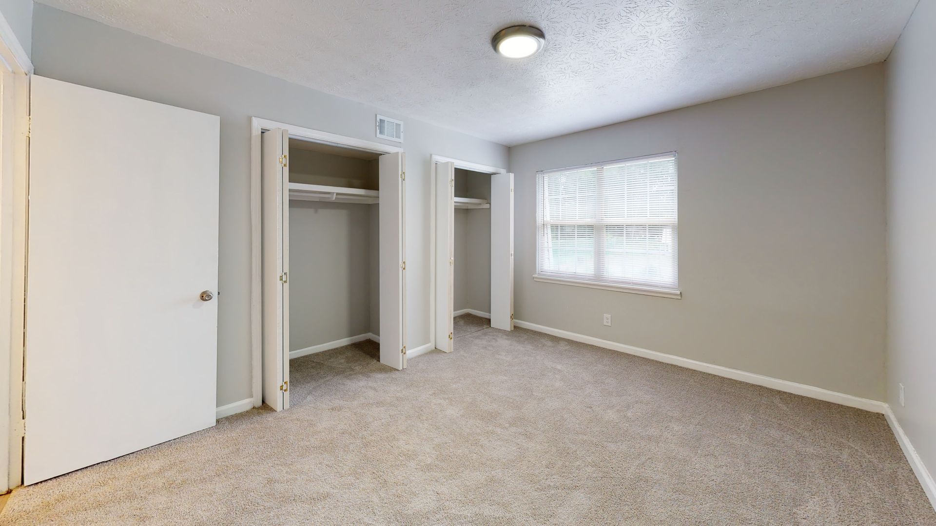 Empty beige bedroom with carpet, open closet doors, and a window with blinds.