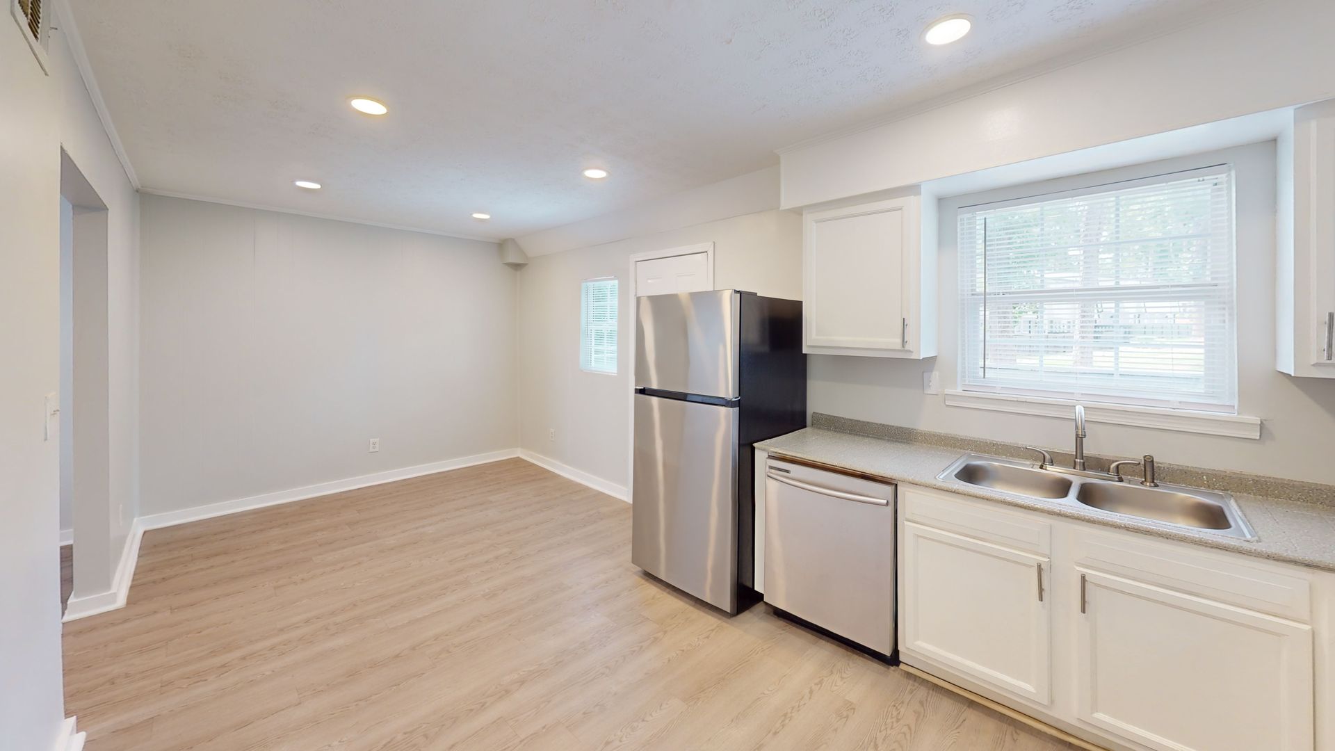 Bright empty kitchen with white cabinets, stainless steel fridge, dishwasher, and light wood floors