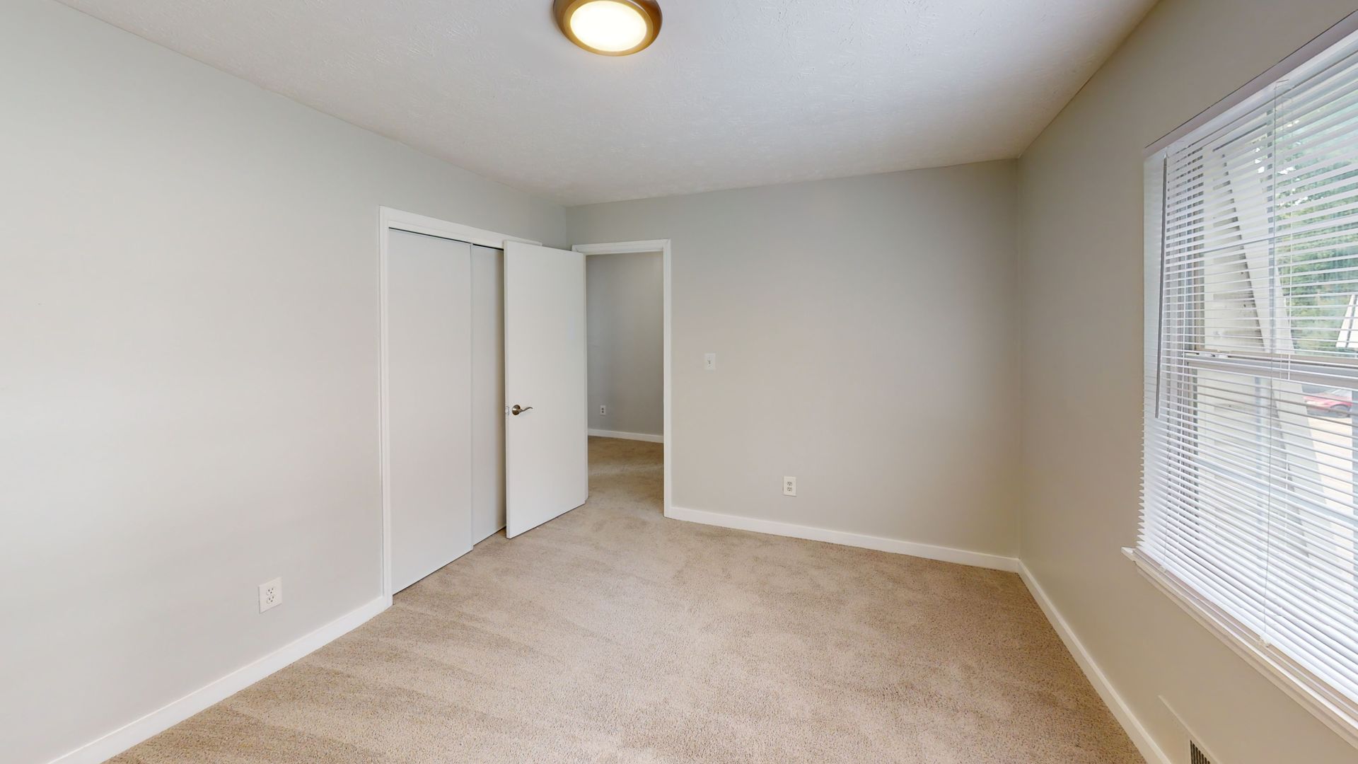 Empty beige bedroom with carpet, white walls, closet door, and a window with blinds.