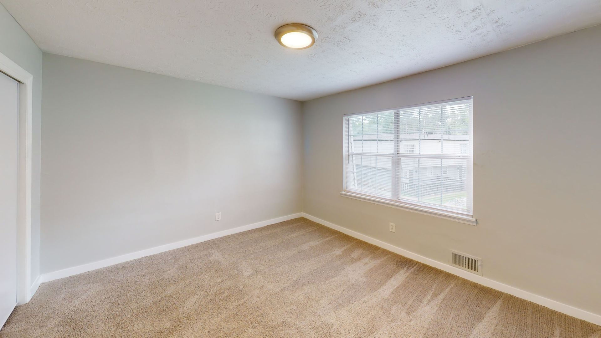 Empty beige bedroom with carpet, white walls, ceiling light, and a window with blinds
