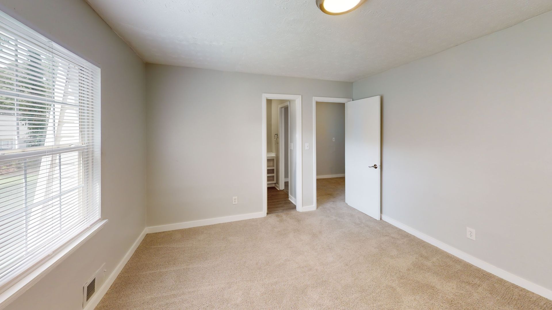 Empty beige bedroom with carpet, window blinds, and an open door to a bathroom hallway