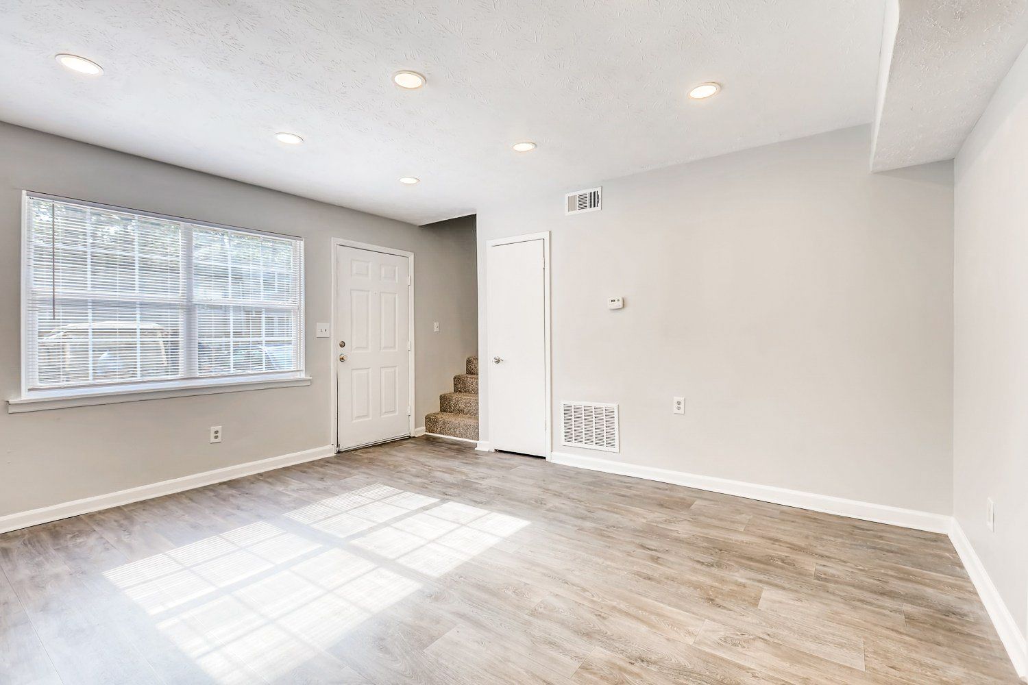 Bright empty living room with white walls, wood floors, window, and stairs in the corner