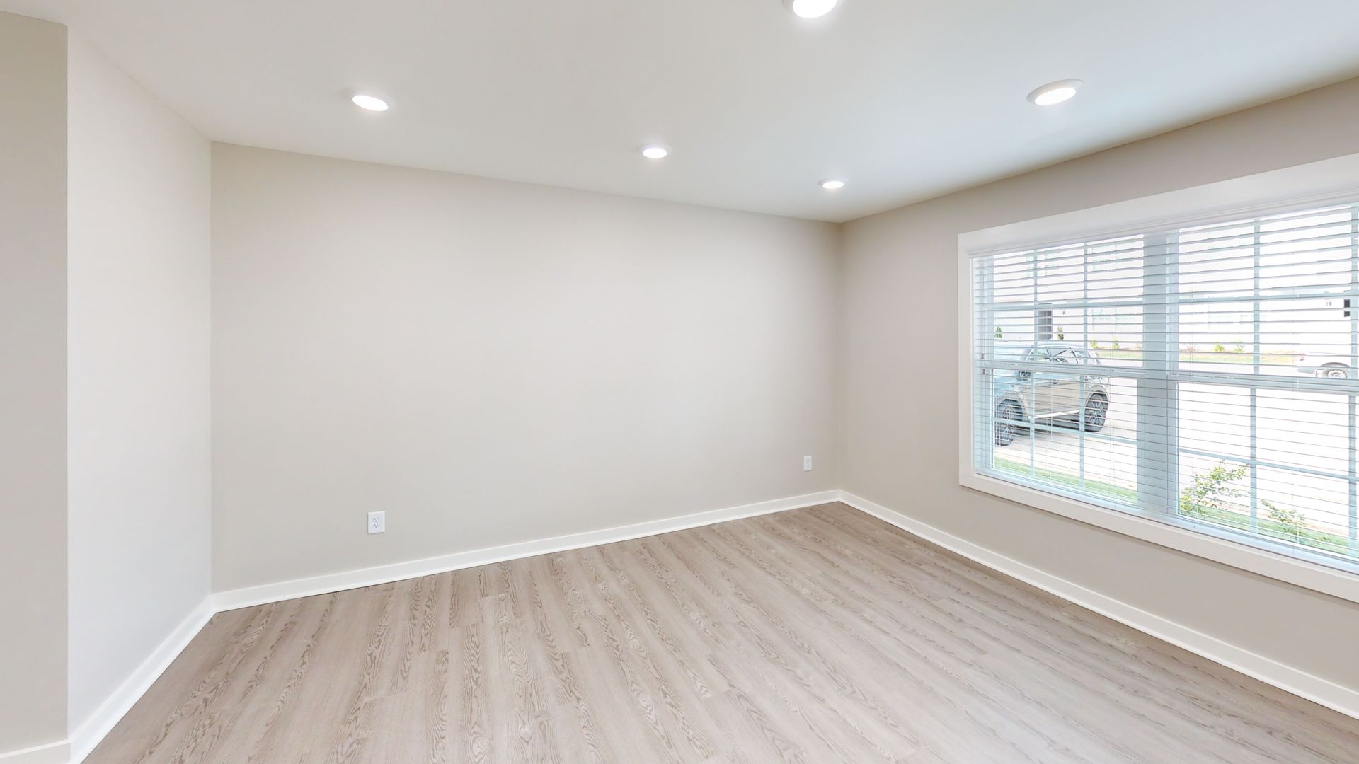 Empty modern room with white walls, light wood floor, recessed lights, and a large window with blinds.