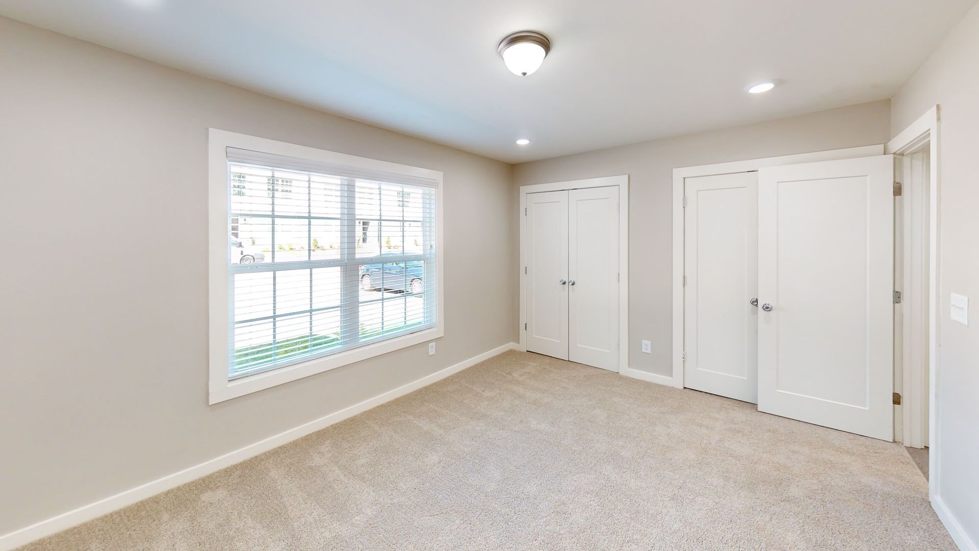 Bright empty bedroom with beige carpet, white walls, a window, and two white closets.