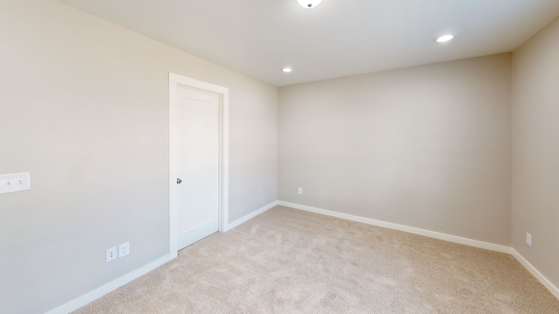 Empty beige bedroom with carpet, white door, and recessed ceiling lights.