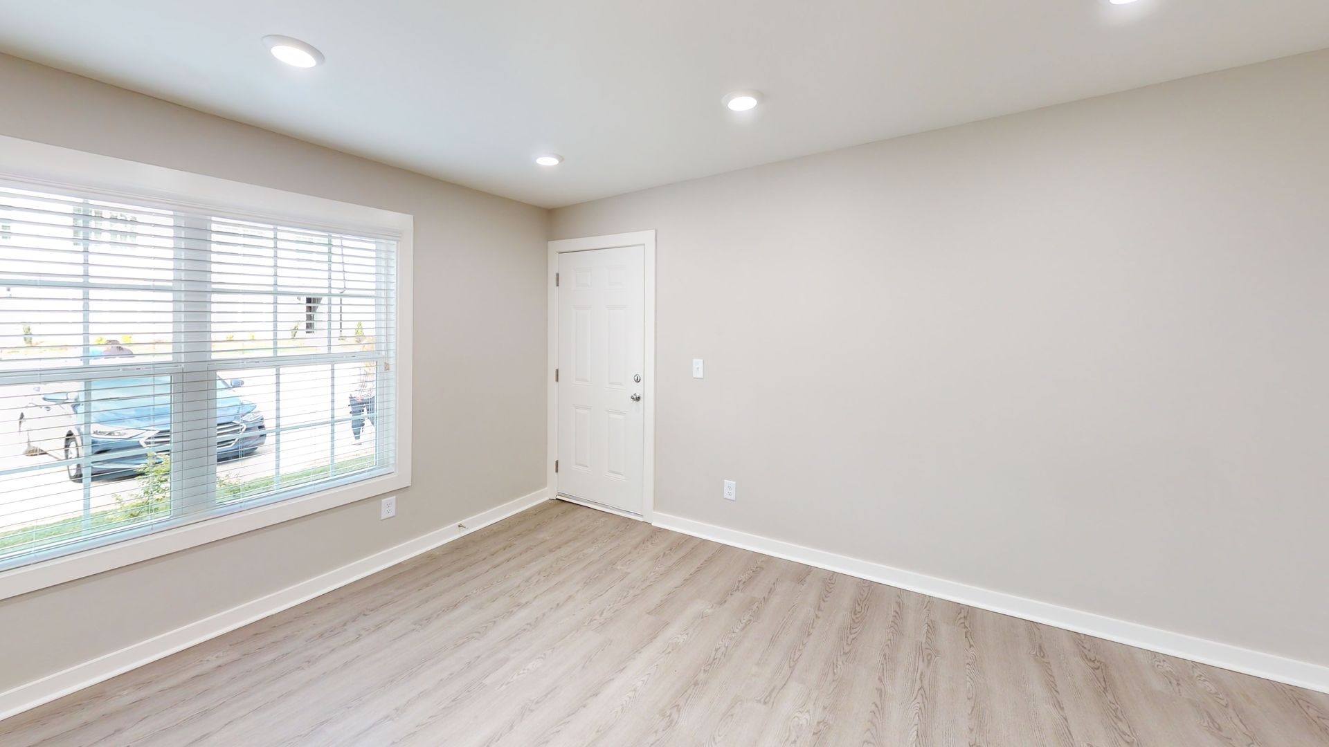 Bright empty room with white walls, light wood floor, recessed lights, and a large window with blinds.